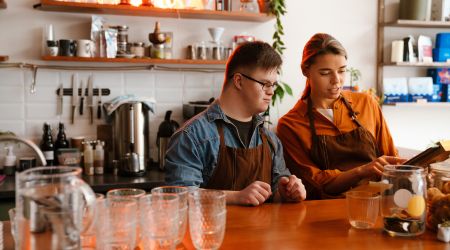 Young boy in glasses working in a cafe looking at a till with a women helping him