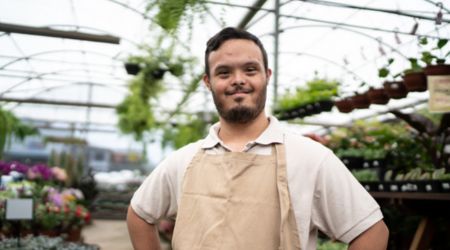 Man stood in a green house with lots of plants around smiling at the camera with hands on hips